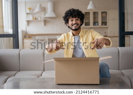Young smiling Indian man unpacking box with present sitting at home. Online shopping, delivery service concept. Successful blogger influencer recording video pointing fingers on carton box, copy space Royalty-Free Stock Photo #2124416771