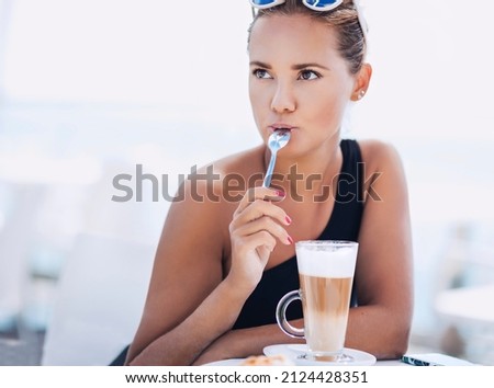 Outdoors lifestyle fashion image of young pretty girl having breakfast. Keeping a small spoon in mouth. Drinking coffee on the beach and enjoying vacation. Wearing stylish sunglasses, swimsuit