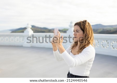 Side view of young female taking picture on modern smartphone while standing on street with white fencing against cloudless sky