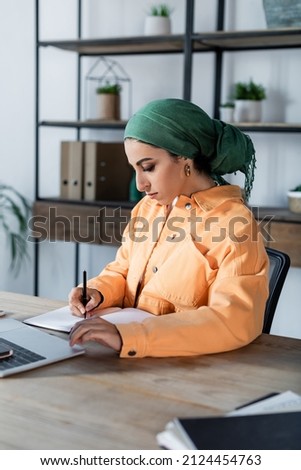 muslim woman in orange jacket and headkerchief writing in notebook while studying at home