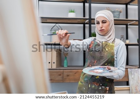 muslim woman with palette and paintbrush near blurred canvas at home