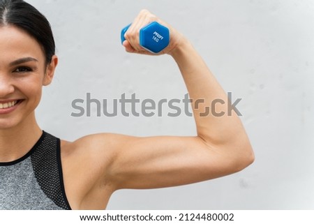 Cropped photo of smiling cheerful female athlete with dumbbell in bent arm looking ahead