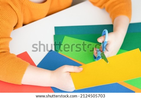 Hands of a child cutting paper with scissors close-up. Blue scissors in left hand, yellow colored paper in right hand. Royalty-Free Stock Photo #2124505703