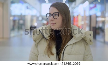 Young woman standing and looking at camera in shopping mall or trade center. Girl portrait in eyeglasses. Shops and showcases, luminous signs. Sale in store.