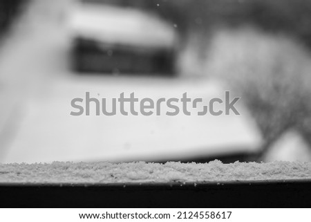 It's snowing. The fence of the terrace is covered with a fine layer of falling snow. In the background an old wooden garage covered with snow. Black and white photograph.
