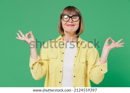Elderly happy woman 50s in glasses yellow shirt hold spreading hands in yoga om aum gesture relax meditate try to calm down isolated on plain green background studio portrait People lifestyle concept