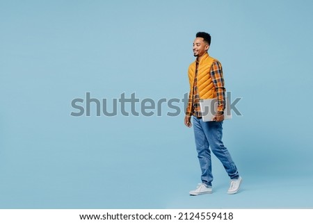 Full size body length fun young black man 20s years old wears yellow waistcoat shirt hold use work on laptop pc computer stepping walking isolated on plain pastel light blue background studio portrait