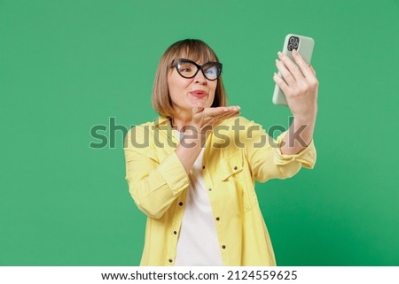 Elderly smiling happy woman 50s in glasses yellow shirt doing selfie shot on mobile cell phone post photo on social network blowing air kiss talk video call isolated on plain green background studio