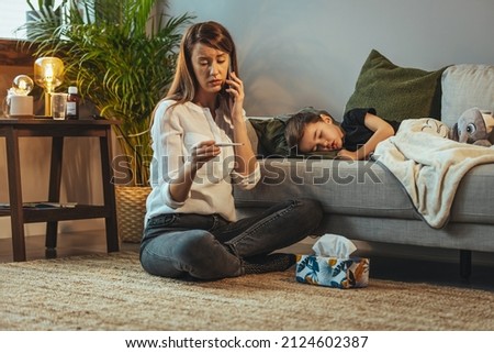 Shot of a mother making a phone call while aiding to her sick young son at home. Mother checking temperature of her sick son. Close up of a mother checking the temperature of her boy  Royalty-Free Stock Photo #2124602387