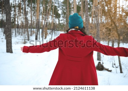 Rear view of unrecognizable woman in green knit woolen hat and bright red warm coat standing with outstretched arms to the side on a snow covered forest, beautiful sunbeams falling through pine trees Royalty-Free Stock Photo #2124611138