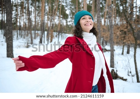 Charming woman in a white sweater, a warm scarf and a bright red woolen coat is whirling, smiling with happiness in a snowy pine forest. Outdoor recreation on a cold snowy winter day. Winter travel Royalty-Free Stock Photo #2124611159
