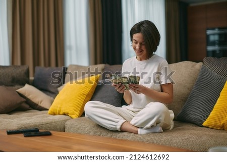 Happy young woman counting cash, enjoying money win, big profit, salary in payday sitting on sofa at home Royalty-Free Stock Photo #2124612692