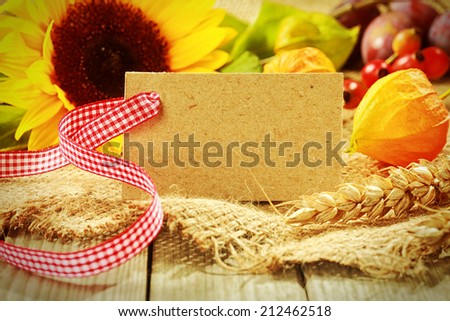 Colorful autumn background with a blank gift tag with red and white ribbon and golden gooseberry, yellow sunflower, rose hips, wheat and berries on coarse textured hessian fabric