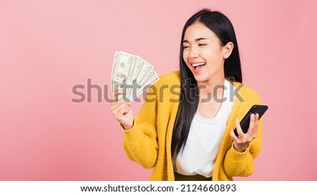 Portrait Asian happy beautiful young woman teen shopper smiling standing excited holding online smart mobile phone and dollar money banknotes on hand in summer, studio shot isolated on pink background
