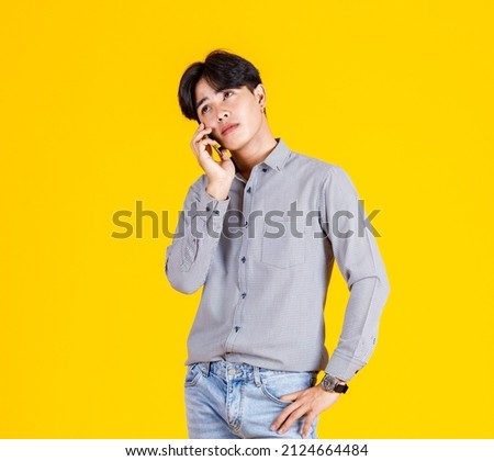 Studio shot of millennial Asian thoughtful doubtful curious male fashion model in stylish fashionable casual outfit standing holding  smartphone on call with friend on yellow background.
