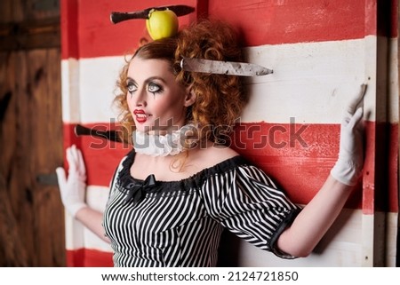 Circus performer girl in a clown costume performs a knife throwing trick. The dark circus concept. Retro style. Royalty-Free Stock Photo #2124721850