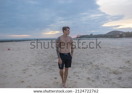 Full height portrait of handsome fit bare-chested young man walking on the beach. Guy with tattoo walking by the sea on the sunset. High quality photo