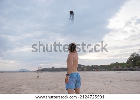Young caucasian fit bare-chested young man walking on the beach and showing on the sky. Balloon in the form of a black octopus flies on the beach. 