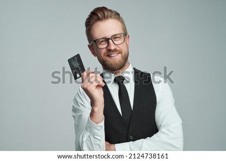 Happy male entrepreneur in glasses showing black plastic card for online purchases while looking at camera against gray background