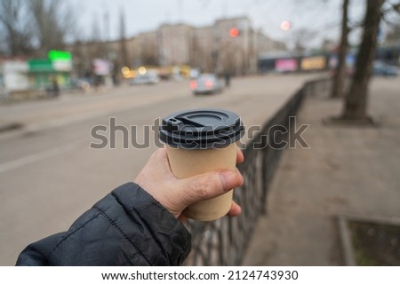 A hand holds a paper cup of coffee against blurry city traffic. A grown man with a hot beverage on the street. Selective focus.