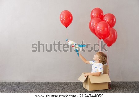 Happy child playing with toy rocket at home. Kid pretend to be astronaut. Boy sitting in cardboard box with red balloons. Imagination and children dream concept
