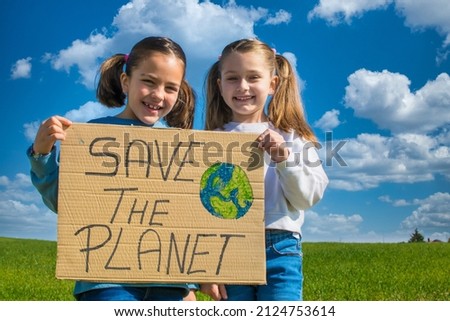 Two Little Girls Holding A Cardboard Sign That Says Save The Planet. They Are In A Green Meadow With A Nice Blue Sky With White Clouds. One Is Blonde And The Other Is Brunette And They Have Pigtails.