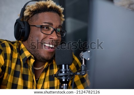 Close-up of an African podcaster blogger smiling while broadcasting his live audio podcast in studio using a microphone, laptop and headphones. Male radio host with glasses, podcast or interview host