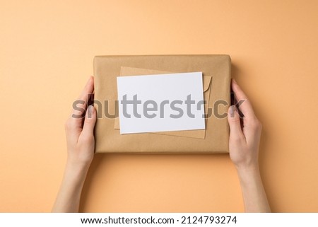 First person top view photo of female hands holding craft paper giftbox with envelope and letter on isolated beige background with empty space