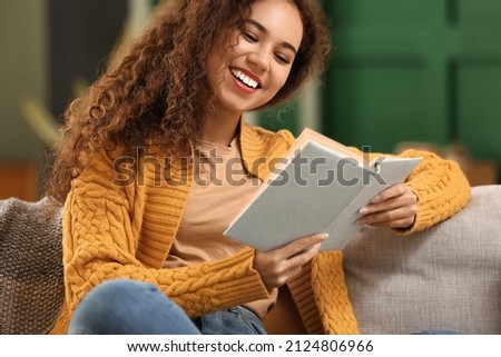 Young African-American woman in yellow cardigan reading book on sofa at home Royalty-Free Stock Photo #2124806966