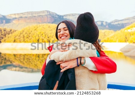 Portrait of two cheerful smiling young female traveler sisters in winter clothes and beanie while standing near mountain lake during winter. Hiking, excursion, traveler concept.