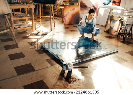 Female painter making a blue painting on a canvas. Creative young woman touching her painting while squatting on the floor in her art studio. Artistic young woman working on a new project. Royalty-Free Stock Photo #2124861305