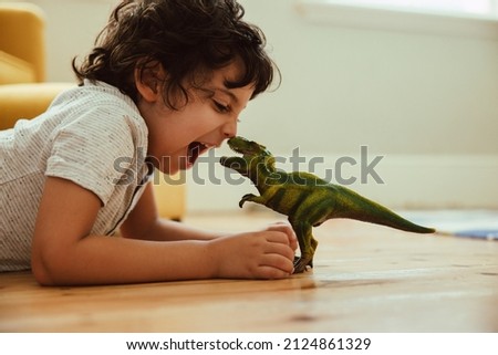 Adventurous young boy imitating a dinosaur toy while lying on the floor in his play area. Creative little boy having fun during playtime at home. Royalty-Free Stock Photo #2124861329