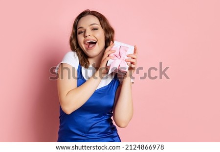 Happy women's day. Excited young beautiful girl with long curly hair holding small gift box isolated on pink background. Concept of emotions, holidays, inspiration, sales, ad. Blue and pink colors