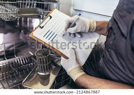 Male Technician Sitting Near Dishwasher Writing On Clipboard In Kitchen, close up. Royalty-Free Stock Photo #2124867887