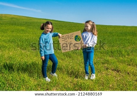 Two Little Girls Holding A Cardboard Sign That Says Save The Planet. They Are In A Green Meadow With A Nice Blue Sky With White Clouds. One Is Blonde And The Other Is Brunette And They Have Pigtails.