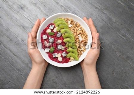Woman holding bowl of granola with pitahaya, kiwi and almonds at grey table, top view