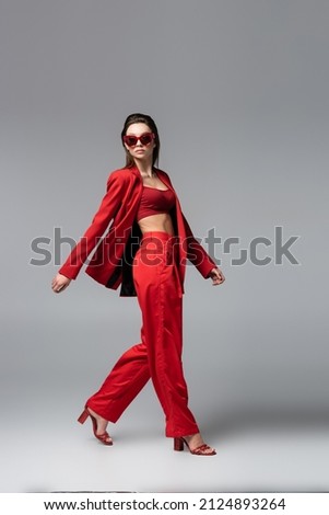 full length of brunette woman in red suit and trendy sunglasses walking on dark grey Royalty-Free Stock Photo #2124893264