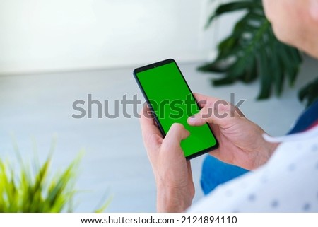 Man holds mobile phone  and playing games online indoors of cozy home. Use green screen for copy space close up. Chroma key mock-up on smartphone in hand.