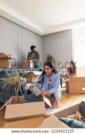 Smiling young woman packing her things in cardboard box as she moves into a new apartment while her roommates talk and help her in the background..