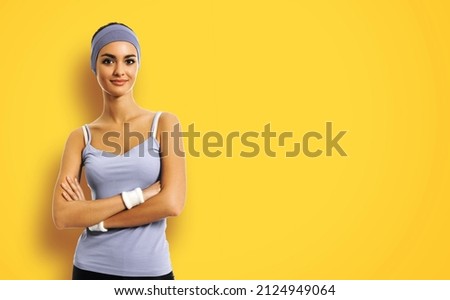 Portrait of smiling sporty brunette woman in grey sportswear, over yellow background. Young female fitness instructor or personal trainer in crossed arms pose at studio.