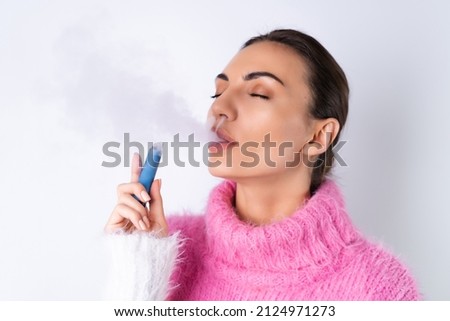A young cheerful girl in a bright colored sweater on a white background enjoys an electronic disposable cigarette with her eyes closed, smokes, releases smoke