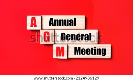 On a bright red background, wooden cubes and blocks with the text AGM Annual General Meeting. Manufacturing of wooden toys. Royalty-Free Stock Photo #2124986129