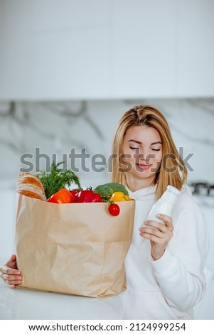 Young mother in white hoodie posing with paper package of fresh vegetables, milk and bread. Delivery food concept. Royalty-Free Stock Photo #2124999254