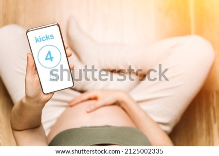 Pregnant woman holding a phone with an app to track baby kicks. Pregnancy apps, gadgets, moves and kicks tracking background Royalty-Free Stock Photo #2125002335