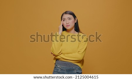 Asian woman feeling worried and emotional on camera in studio. Negative person showing anxious emotion and frustration, worrying about problems, standing over yellow background. Royalty-Free Stock Photo #2125004615