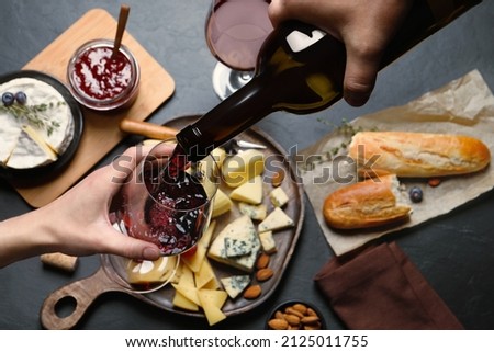 Man pouring red wine from bottle into glass over black table with snacks, top view