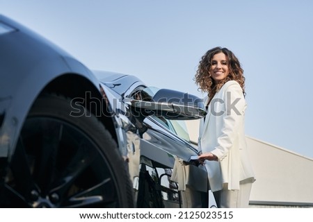 Bottom view of happy smiling business girl with curly hair standing outside car and easily opening door of her auto. Beautiful woman holding handle on car door on the background of blue sky.