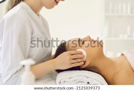 Female client enjoying cosmetic spa facial treatment at beauty parlour. Professional beautician cleaning young girl's skin with cotton discs. Side profile view of woman's head on soft white towel Royalty-Free Stock Photo #2125039373