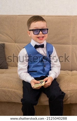 A boy in a blue vest and dark glasses holds a wad of money in his hands while sitting on the sofa. A young businessman