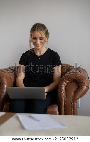 Young woman working at home with laptop on a brown arm chair with a white background. Home office concept. Gray notebook for working. Home office concept. High quality photo
